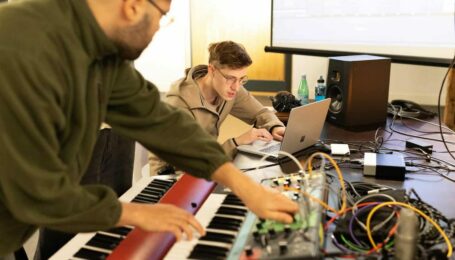 In de voorgrond speelt een man op een keyboard, en achter hem werkt iemand op een laptop.